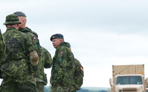 A group of military personnel in green camouflage uniforms are gathered on a grassy area beside a gravel road, appearing to be in discussion or receiving a briefing. Several soldiers display flag patches on their shoulders consistent with Danish military insignia. In the background, a convoy of tan/desert-colored military vehicles, including a heavy transport truck, is visible on the road.