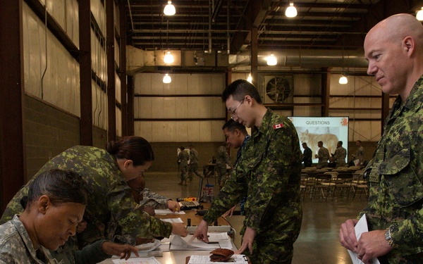 Military personnel from the United States Army and Canadian Armed Forces are gathered inside a large hangar or warehouse facility, engaged in what appears to be a processing or in-processing administrative event. A US Army soldier seated at a table is reviewing and highlighting documents, while Canadian soldiers in CADPAT camouflage uniforms stand at the table presenting paperwork. A presentation screen displaying 'QUESTIONS' is visible in the background, suggesting a briefing recently concluded.