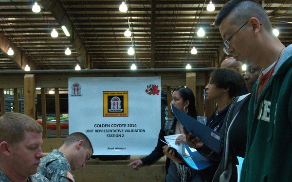 The image shows a military administrative processing event identified as 'Golden Coyote 2014 Unit Representative Validation Station 2' at a headquarters location. Two soldiers in Army Combat Uniforms (ACU) are seated at a table processing paperwork for a line of civilians and personnel. The setting appears to be a large indoor facility such as a warehouse or armory.