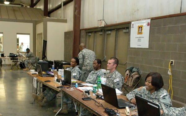 Military personnel in camouflage uniforms are gathered in a large indoor facility, likely a warehouse or armory. Several soldiers are seated at long folding tables equipped with laptops and paperwork, while two others stand facing them, suggesting a briefing, processing, or administrative operation. The setting appears to be a mobilization or readiness station.
