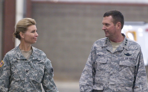 Two U.S. military officers are walking together inside what appears to be a military hangar or large indoor facility. The officer on the left wears a U.S. Army uniform with the name tape 'KUHN' and a colonel's eagle insignia, while the officer on the right wears a U.S. Air Force uniform with the name tape 'LEMOGYEL' and visible general officer stars. Additional personnel are visible in the background.