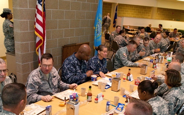 Military personnel in various branch uniforms are gathered in what appears to be a military dining facility (DFAC/mess hall). Soldiers primarily in Army ACU (Army Combat Uniform) pattern camouflage, along with at least two individuals in Navy uniforms, are seated at long tables sharing a meal. The setting appears to be an official or semi-formal dining event, possibly a command luncheon or unit gathering.