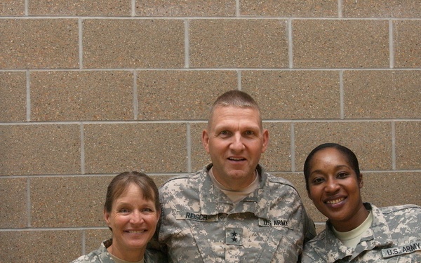 Three U.S. Army soldiers in ACU (Army Combat Uniform) digital camouflage are posing together for a photo in front of a brick wall. The center figure appears to be a general officer based on the visible star insignia on the chest. They are flanked by the U.S. flag on the left and what appears to be the South Dakota State flag on the right.