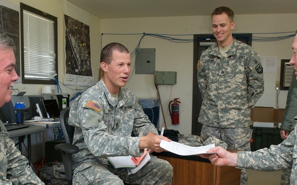 Four U.S. Army soldiers in ACU (Army Combat Uniform) digital camouflage are gathered in what appears to be a military office or operations room. Three soldiers are seated while one stands in the background, and the central focus is on a document being passed or reviewed between two of the seated soldiers. The setting suggests a briefing, administrative review, or planning session.