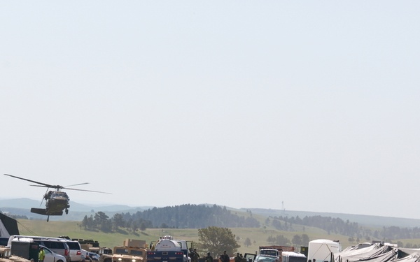 A military field base or forward operating base is depicted in an open grassy area, likely during a training exercise. Multiple military personnel are visible moving along a dirt road flanked by military tents, vehicles, and equipment. A helicopter, appearing to be a UH-60 Black Hawk, is visible in flight on the left side of the image.