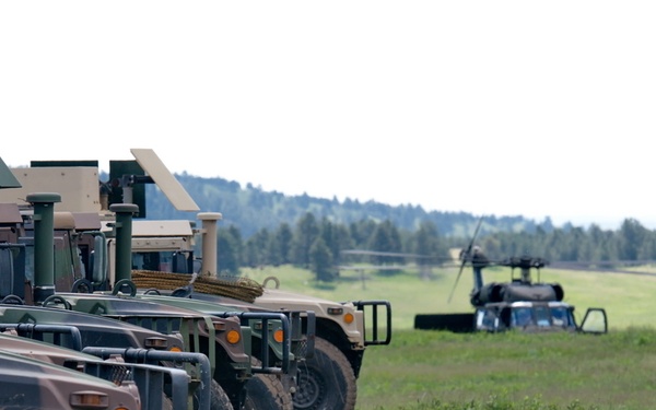 A U.S. Army soldier in ACU (Army Combat Uniform) walks alongside a row of parked military Humvees (HMMWVs) on an open grassy field. The vehicles are lined up in formation, some equipped with mounted weapon systems or communications equipment. In the background, what appears to be a grounded helicopter is visible among trees on a rolling landscape.