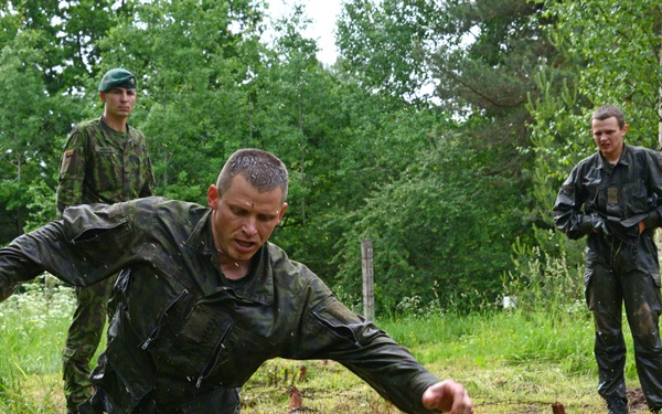 Three military personnel are visible in an outdoor field training environment surrounded by trees and vegetation. The central figure, wearing camouflage fatigues, is actively running or stumbling through muddy terrain with water and mud splashing around his feet. Two other soldiers stand in the background observing, one wearing a green beret.