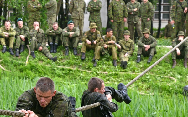Two soldiers in military uniforms are traversing a rope suspended over a marshy, reed-filled water obstacle as part of a military training exercise. A large group of uniformed soldiers watches from an embankment in the background. One soldier appears soaking wet, suggesting he may have already fallen into the water.