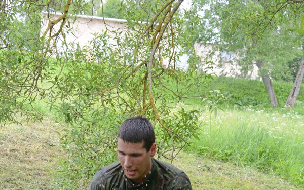 A young man in olive green military-style clothing is navigating what appears to be a physical training obstacle course. He is soaked and covered in mud, actively moving through a muddy pit area with wooden stakes and a tire obstacle. The action shot captures splashing muddy water, suggesting intense physical exertion.