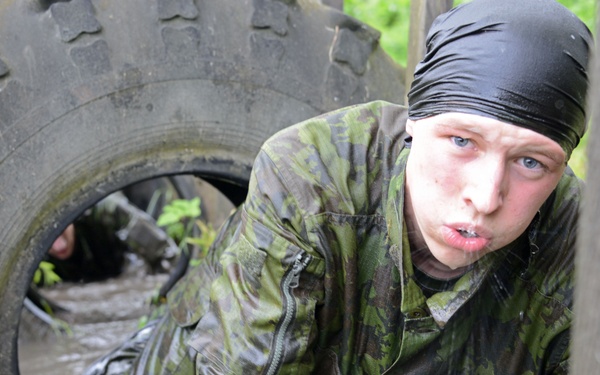 A person wearing camouflage military fatigues and a black head covering is crawling through a muddy water obstacle course, emerging from beneath a large tire. The individual appears physically exerted and is soaking wet with mud. A second person is partially visible in the background navigating the same obstacle.