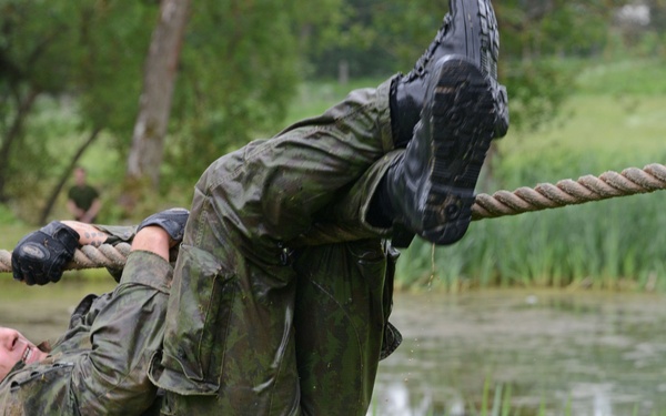 A soldier in wet camouflage uniform is traversing a rope obstacle over a body of water, hanging upside-down or at an inverted angle while gripping the rope with both hands and legs. The setting appears to be a military training course or obstacle course in a natural outdoor environment. The uniform and gear are visibly soaked and muddy, indicating the exercise has been physically demanding.