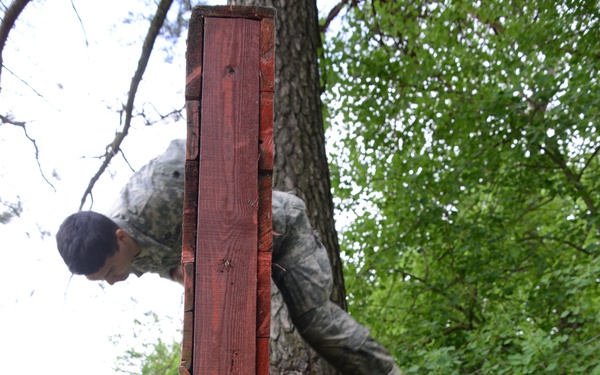 Two individuals in military camouflage uniforms are navigating a wooden obstacle course structure in a wooded outdoor setting. One person is climbing over the top of a tall red-painted wooden wall obstacle while the other stands below, appearing to assist or observe. The structure consists of vertical planks supported by diagonal log braces, typical of military training course obstacles.