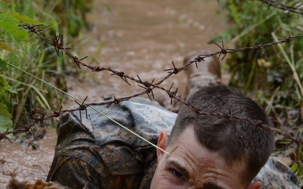 A soldier in camouflage uniform is crawling through a flooded, muddy trench under barbed wire obstacles. The individual appears to be completing a military obstacle course or combat training exercise, with their face barely above the muddy water. The expression on the soldier's face conveys intense physical exertion and determination.