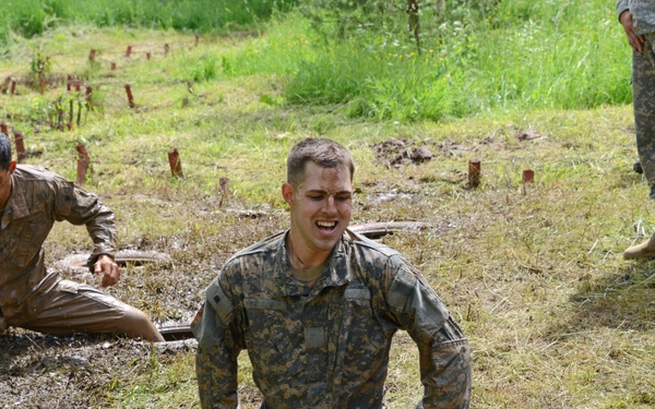 Military personnel are participating in a physically demanding obstacle course or field training exercise in a muddy outdoor environment. Two soldiers in the foreground are covered in mud and appear to be emerging from or navigating through ground-level obstacles, including circular hatch openings in the ground. Additional uniformed personnel are visible observing or supervising the activity.