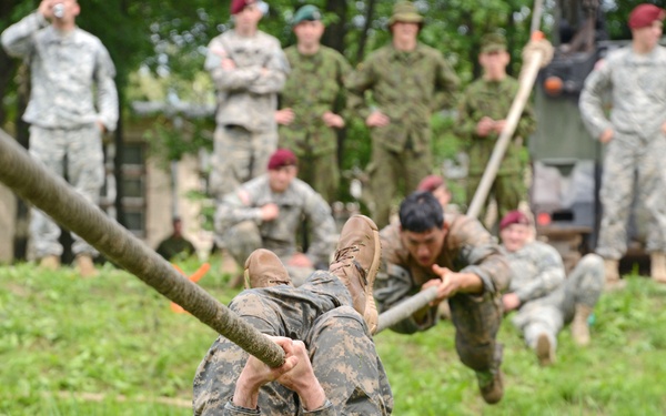 Military personnel are navigating a rope obstacle course over a grassy/marshy area. The foreground soldier is hanging upside down from a horizontal rope, gripping it with hands and feet, while a second soldier follows behind on the same rope. A large group of observers in various military uniforms watches from the background.