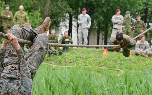 Military personnel are participating in a rope obstacle course training exercise outdoors. A soldier in camouflage uniform is hanging upside down from a rope in the foreground, while another soldier traverses a rope in the background. A large group of multinational military observers in various uniforms watch from the sidelines.