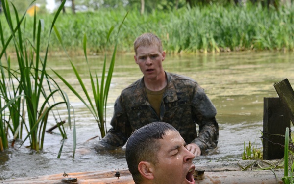 Two military personnel in camouflage uniforms are navigating a water obstacle course, likely during military training. The foreground soldier is submerged up to his neck, mouth open wide, pushing under or through a log barrier in murky water. A second soldier follows behind in the background, both appearing physically exerted.