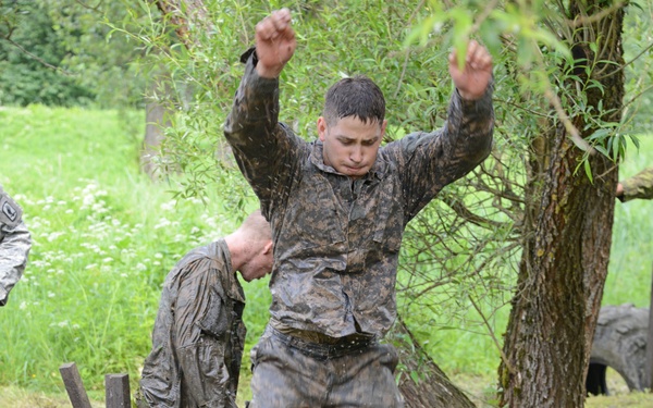 Two individuals in camouflage military uniforms, covered in mud and water, are navigating a military-style obstacle course. The foreground subject has both arms raised while jumping through or over a series of tires embedded in muddy ground with wooden stakes. The setting appears to be an outdoor training area with green vegetation and trees in the background.
