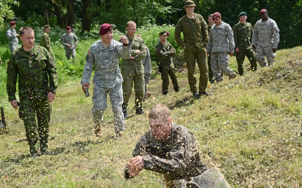 A group of military personnel from multiple nations are gathered on a grassy hillside, observing a demonstration or training exercise. One soldier in the foreground is crouched low to the ground near what appears to be a small controlled detonation or explosive device, with debris visibly spraying outward. The group includes soldiers in US Army ACU pattern uniforms, olive drab uniforms, and personnel wearing red and maroon berets.