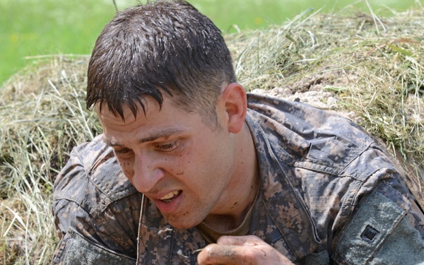 A soldier in digital camouflage uniform is crawling through a narrow mud tunnel or low-crawl obstacle, emerging from the opening covered in mud and dirt. The individual appears to be undergoing military training, specifically a combat or obstacle course exercise. The expression on the soldier's face suggests physical exertion and determination.