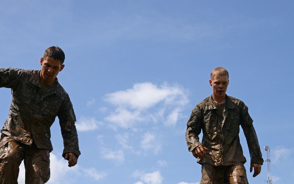 Two individuals in military-style camouflage uniforms, heavily covered in mud, are navigating what appears to be an elevated obstacle course structure made of green metal pipes. The low-angle shot captures them balancing on top of the structure against a partly cloudy blue sky. This appears to be a military training exercise or obstacle course event.
