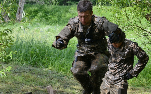 Two individuals in camouflage military uniforms, visibly covered in mud, are navigating an outdoor obstacle course. One person is mid-jump over an obstacle consisting of wooden stakes, tires, and barbed wire, while the second person stands nearby observing or preparing. The setting is a wooded, grassy area suggesting a military training exercise.