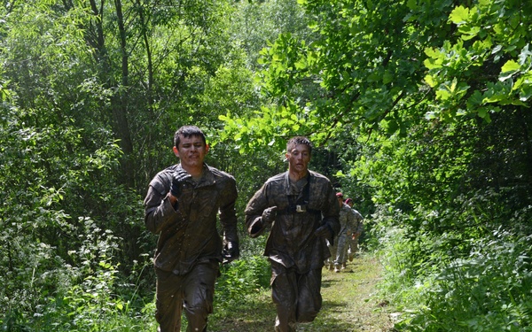 A group of individuals in mud-covered military-style uniforms are running along a narrow dirt trail through a dense, green forested area. Two individuals lead the group in the foreground, both visibly covered in mud, suggesting participation in a military training exercise or obstacle course event. Additional participants are visible in the background following the same trail.