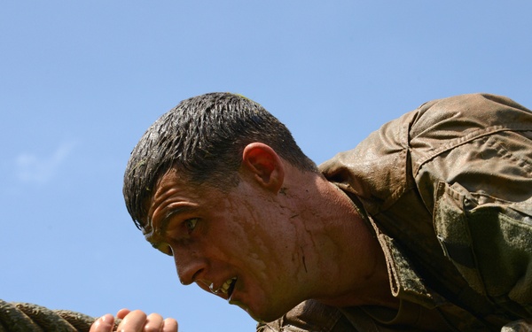 A male individual wearing mud-soaked military camouflage clothing is gripping a thick rope with both hands, appearing to pull or climb it with intense physical effort. His face and uniform are visibly wet and covered in mud, suggesting participation in a military obstacle course or physical training exercise. The image is taken from a low angle against a clear blue sky, emphasizing the strain and determination on his face.