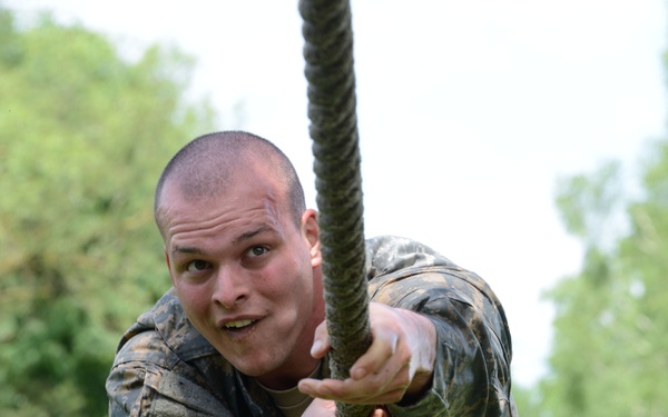 A young male in military camouflage uniform is climbing or gripping a thick rope, displaying visible physical exertion. His uniform appears muddy or worn, suggesting an outdoor training exercise or obstacle course. The background shows green foliage and an overcast sky.