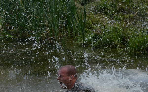 A bald man is captured mid-splash as he moves forcefully through a muddy, shallow body of water. He appears to be shouting or gasping, with water erupting dramatically around him. The setting appears to be a natural waterway with reeds and green vegetation visible in the background.