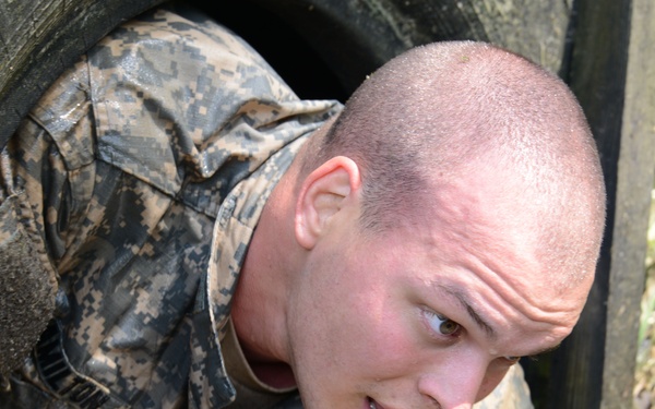 A young male soldier in US Army ACU (Army Combat Uniform) digital camouflage is crawling through a large tire obstacle. His uniform is visibly muddy and wet, indicating an outdoor physical training or obstacle course exercise. His facial expression shows concentration and physical exertion.