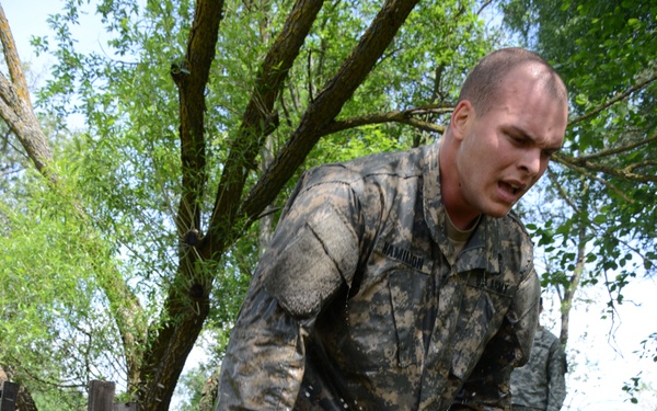 A U.S. Army soldier, identified by name tape as 'Hamilton,' is captured mid-stride running through a muddy water obstacle during what appears to be a military training exercise or obstacle course. His uniform (ACU pattern) is heavily soaked and caked with mud, indicating prolonged physical exertion through wet terrain. A second soldier in similar uniform is visible in the background observing or participating in the same exercise.