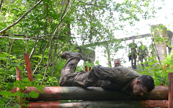 A mud-covered soldier or trainee is crawling over a low wooden log obstacle in a forested outdoor training course. The individual is prone on the logs, covered in mud, suggesting an intense physical endurance exercise. In the background, other uniformed personnel are visible observing or participating in the course near what appears to be a ruined structure.