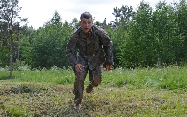 A soldier in camouflage uniform is running across a muddy, grassy field during what appears to be a military training exercise. The uniform and boots are visibly covered in mud, suggesting the soldier has been engaged in a physically demanding outdoor course. The setting is a rural or forested training area with trees and utility poles visible in the background.
