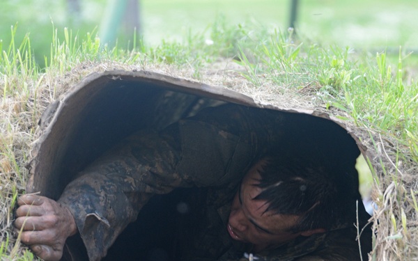 A person wearing camouflage military uniform is crawling through a narrow underground tunnel or culvert pipe, covered in mud. The individual appears to be completing a military-style obstacle course or training exercise. Both hands are visible gripping the tunnel edges as they emerge from the opening.
