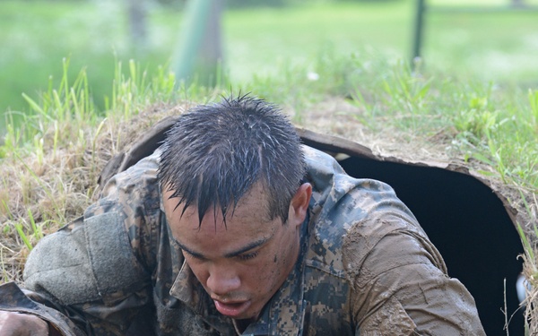 A soldier in digital camouflage uniform is crawling out of a tunnel or underground passage during what appears to be a military obstacle course or training exercise. The individual is heavily covered in mud, with wet hair and soiled uniform, indicating intense physical exertion in wet conditions. The setting is an outdoor grassy field with a dirt tunnel opening visible in the foreground.