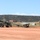 Military personnel are operating what appears to be a UAV (unmanned aerial vehicle) catapult launch system on a dirt surface at a military installation. A small fixed-wing drone is positioned on the launch rail of a wheeled, olive-drab colored launcher. The setting appears to be a high-desert environment with scrub-covered mesas visible in the background.