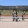 Four military personnel in U.S. Army camouflage uniforms are handling a small unmanned aerial vehicle (UAV) on a gravel surface. One soldier is positioned at the center of the aircraft appearing to guide or position it, while the others walk alongside. The setting appears to be a remote airstrip or training area with mesa terrain and sparse vegetation in the background.