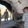 A group of military personnel in US Army ACU (Army Combat Uniform) camouflage are gathered inside a tent or temporary shelter examining an unmanned aerial vehicle (UAV/drone). The aircraft, marked with the number '240G', appears to be a Shadow RQ-7 or similar tactical UAV used for reconnaissance. A military HMMWV (Humvee) is visible in the background outside the shelter.