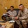 Two men are seated on leather sofas in a plywood-walled room, engaged in what appears to be a meeting or interview. One man wears US Marine Corps desert MARPAT camouflage uniform with a gold oak leaf insignia (Major rank), while the other is in civilian clothing and appears to be taking notes. A coffee table in front of them holds snacks, candies, notebooks, and pens.