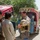 A Western man wearing a body armor vest and carrying a military-style helmet is engaged in a conversation with two local men dressed in traditional shalwar kameez. The scene takes place outdoors on a dusty road beside colorfully decorated auto-rickshaws (tuk-tuks), typical of South or Central Asian regions. The Western man appears relaxed and is smiling during the interaction.