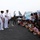 Two naval officers in white dress uniforms are addressing a large group of Korean civilians, including many children, on the flight deck of an aircraft carrier. A military jet aircraft is visible in the background, along with additional naval personnel. The banner held by visitors contains Korean text, suggesting this is an organized public tour or community outreach event.