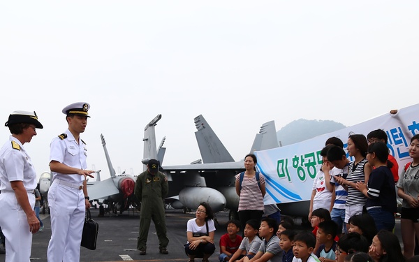 Two naval officers in white dress uniforms are addressing a large group of Korean civilians, including many children, on the flight deck of an aircraft carrier. A military jet aircraft is visible in the background, along with additional naval personnel. The banner held by visitors contains Korean text, suggesting this is an organized public tour or community outreach event.