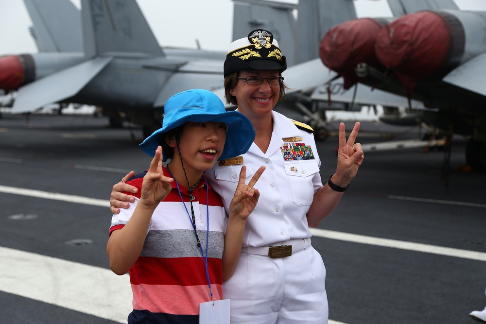 A naval officer in white dress uniform poses with a young visitor on the flight deck of an aircraft carrier. Both individuals are making peace/victory hand signs for the photo. Military aircraft with red protective covers on their engines are visible in the background.