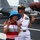 A naval officer in white dress uniform poses with a young visitor on the flight deck of an aircraft carrier. Both individuals are making peace/victory hand signs for the photo. Military aircraft with red protective covers on their engines are visible in the background.