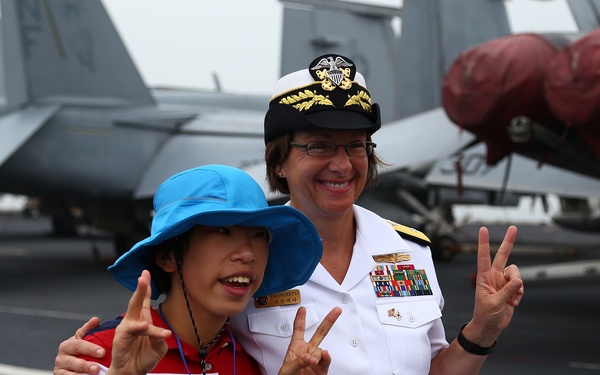 A naval officer in white dress uniform poses with a young visitor on the flight deck of an aircraft carrier. Both individuals are making peace/victory hand signs for the photo. Military aircraft with red protective covers on their engines are visible in the background.