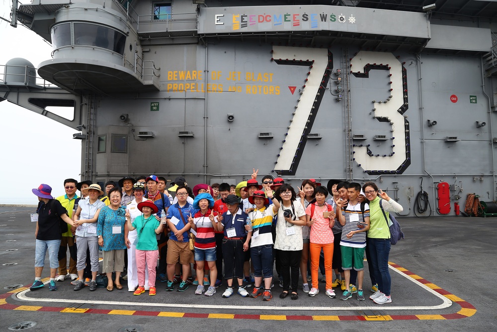 A large group of civilian visitors, appearing to be of East Asian origin, pose for a group photo on the flight deck of USS George Washington (CVN-73), a US Navy aircraft carrier. The ship's island superstructure is visible behind them, displaying the hull number '73' and a safety warning reading 'BEWARE OF JET BLAST PROPELLERS AND ROTORS'. The visitors are wearing colorful casual clothing and many are making peace/victory hand signs.