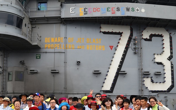 A large group of civilian visitors, appearing to be of East Asian origin, pose for a group photo on the flight deck of USS George Washington (CVN-73), a US Navy aircraft carrier. The ship's island superstructure is visible behind them, displaying the hull number '73' and a safety warning reading 'BEWARE OF JET BLAST PROPELLERS AND ROTORS'. The visitors are wearing colorful casual clothing and many are making peace/victory hand signs.