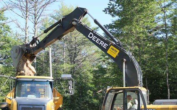 A construction site in a wooded area features a John Deere excavator (model 700D) loading dirt or soil into a John Deere articulated dump truck. A worker in a green safety vest and hard hat stands in the foreground observing the operation. The equipment appears to be rented, as indicated by a visible Hertz Equipment rental sticker on the excavator boom.