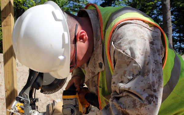 A worker wearing US Marine Corps MARPAT camouflage uniform, a white hard hat, and a high-visibility green and orange safety vest is operating a power saw on lumber at an outdoor construction site. The individual appears to be cutting or measuring a wooden beam using a yellow DeWalt circular saw. A miter saw is also visible in the background on the same work surface.